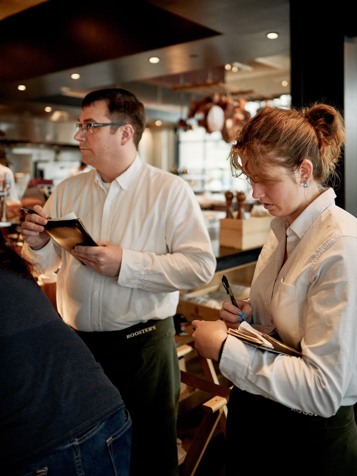 Two servers taking pre-shift notes by the window, copper pans behind them